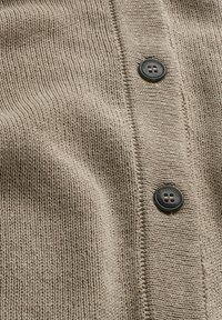 Close-up of beige knitted fabric with two black buttons sewn on a vertical strip, buttons engraved with "ARME" and "ANGELS."
