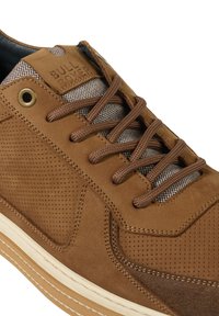 Brown suede shoe with perforated detailing, brown laces, and a checkered textile tongue. Features a cream rubber sole and embossed logo.
