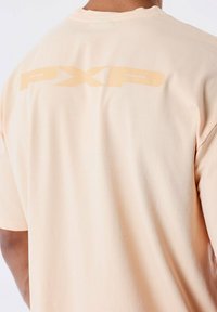 Camiseta de melocotón claro hecha de una tela suave, con un logo simple en un tono más oscuro en la parte de atrás. Manga corta y corte estándar.