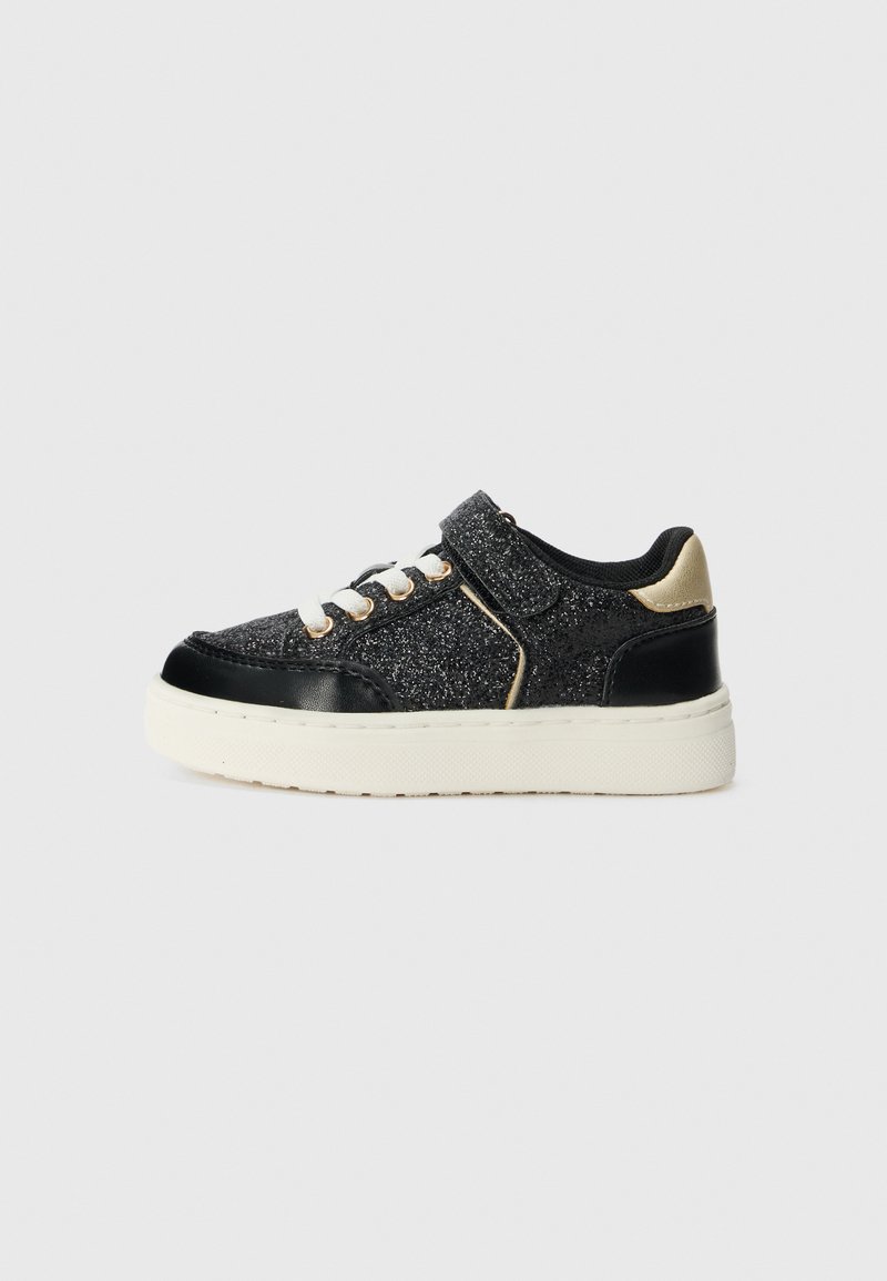 Sneaker nera con tomaia glitterata, accenti in pelle verniciata nera, linguetta beige, lacci bianchi e cinturino in Velcro. Suola bianca spessa.