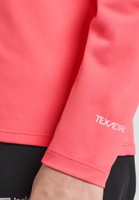 Camicia a maniche lunghe di un rosa corallo realizzata in tessuto testurizzato. Presenta un logo bianco "TEXADRI" sulla manica. Vista ravvicinata del braccio e del polsino.