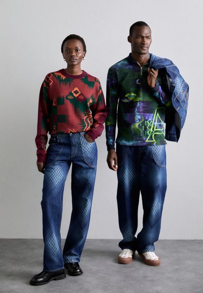 Colorful patterned sweater in red, green, paired with blue jeans featuring diamond patterns. Man wears a dark green patterned top and denim jacket.