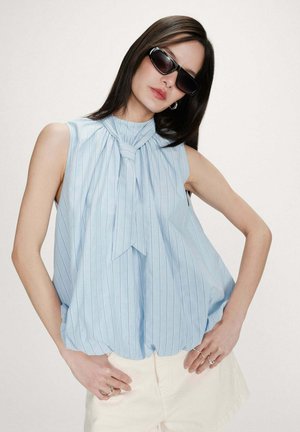 Femme aux cheveux bruns raides portant des lunettes de soleil noires, un haut sans manches bleu clair à rayures avec un nœud au col, et un pantalon blanc, posant les mains sur les hanches.