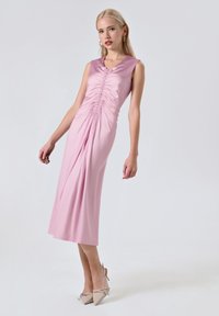 Rosa ärmelloses Kleid mit einer gerafften Textur vorne, das bis zur Mitte der Wade fließt, kombiniert mit hellen Absatzschuhen.