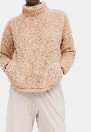 Person iført en beige, tekstureret fleecebluse med høj krave og forlommer, kombineret med lyse bukser.