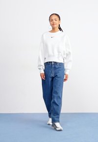 Sudadera de cuello redondo, gris claro con un logo negro, combinada con jeans azules holgados y zapatillas blancas con detalles grises. Pendientes circulares visibles.