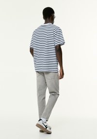Mann mit einem gestreiften T-Shirt in Weiß und Marineblau, hellgrauen Jeans und weißen Turnschuhen, der sich vor einem schlichten weißen Hintergrund abwendet.
