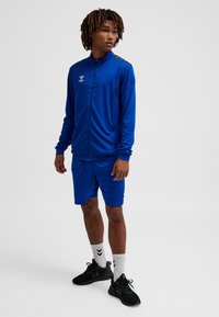 Blauer Sportanzug aus leichtem Stoff, bestehend aus einer durchgehenden Reißverschlussjacke mit Logo und passenden Shorts, kombiniert mit schwarzen Turnschuhen.