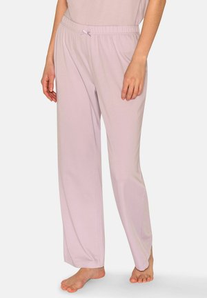 Frau trägt lockere, hellrosa Schlafanzughose mit elastischem Bund und kleinem Schleifendetail, steht barfuß vor einfachem Hintergrund.