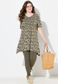 Tunique vert olive avec un motif floral blanc, encolure smockée et manches courtes, associée à des leggings verts olive et un sac fourre-tout tressé beige.