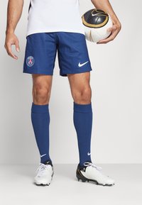 Blauwe voetbalkorte broek van Nike met het embleem van Parijs, gecombineerd met blauwe kniekousen en witte voetbalschoenen met zwarte accenten. Houdt een gemusterde bal vast.
