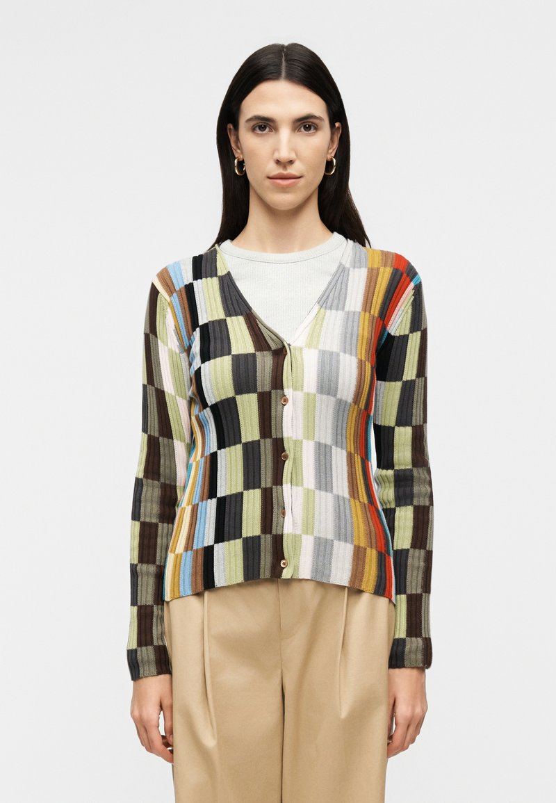 Γυναίκα με μακριά σκουρόχρωμα μαλλιά που φοράει χρυσούς κρίκους στα αυτιά, πολύχρωμη ριγέ ζακέτα με κουμπιά και μπεζ παντελόνι ψηλής μέσης, στέκεται μπροστά.