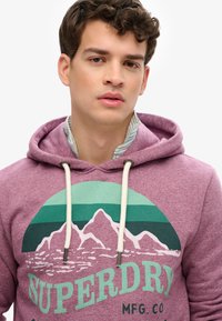 Fioletowa bluza z kapturem z grafika zielonej góry oraz logo "SUPERDRY", z białymi sznurkami i miękką teksturą. Nałożona na paskowaną koszulę.