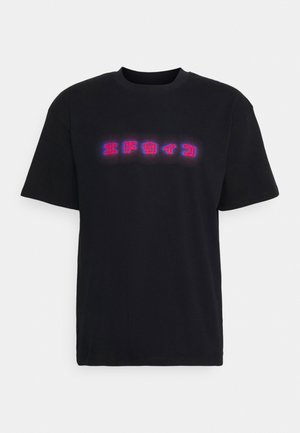 Jeune personne portant un T-shirt noir avec un texte rouge et bleu lumineux en caractères japonais, tenant un sweat rouge plié.