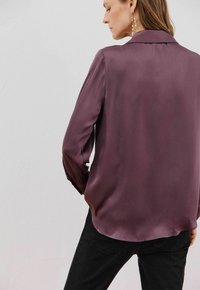 Camicia di seta prugna a maniche lunghe con colletto e texture liscia. Presenta una lieve lucentezza e un orlo dritto, abbinata a jeans neri.