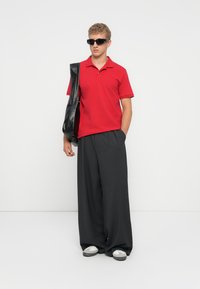 Rotes Polohemd, schwarze weite Hose und schwarze Sonnenbrille. Hält eine schwarze Lederhandtasche. Schuhe sind weiß mit schwarzen Akzenten.