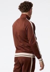 Hnědá sportovní bunda s vysokým límcem, kontrastní bílé pruhy podél rukávů a boků a žebrovaný lem a manžety. Hladká textura látky.