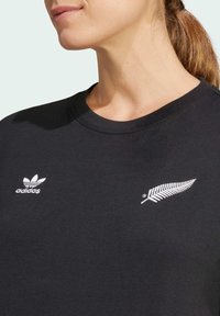 Svart bomull t-shirt med vit broderad Adidas-logotyp och design av New Zealand fern på vänster sida samt rund hals.