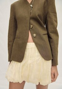 Olive green blazer with two round buttons, paired with a pleated, cream-colored mini skirt. The outfit features a relaxed fit and smooth texture.