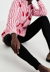 Chemise rayée rouge et blanche avec appliqués floraux, associée à des leggings en daim noir et des chaussures à imprimé léopard. Gros plan sur une pose assise.