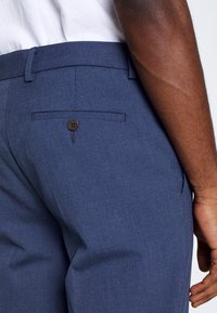 Mörkblå byxor i ett strukturerat tyg, med en bakficka som stängs med en brun knapp och en slim, rak bendesign.