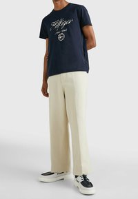 Marinblå bomullströja med vit skriptlogotyp, tillsammans med gräddfärgade vida byxor och svart-vita sneakers.