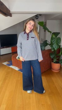 Young woman standing indoors wearing a striped long-sleeve shirt, wide-leg jeans, and silver shoes beside a large potted plant and sofa.