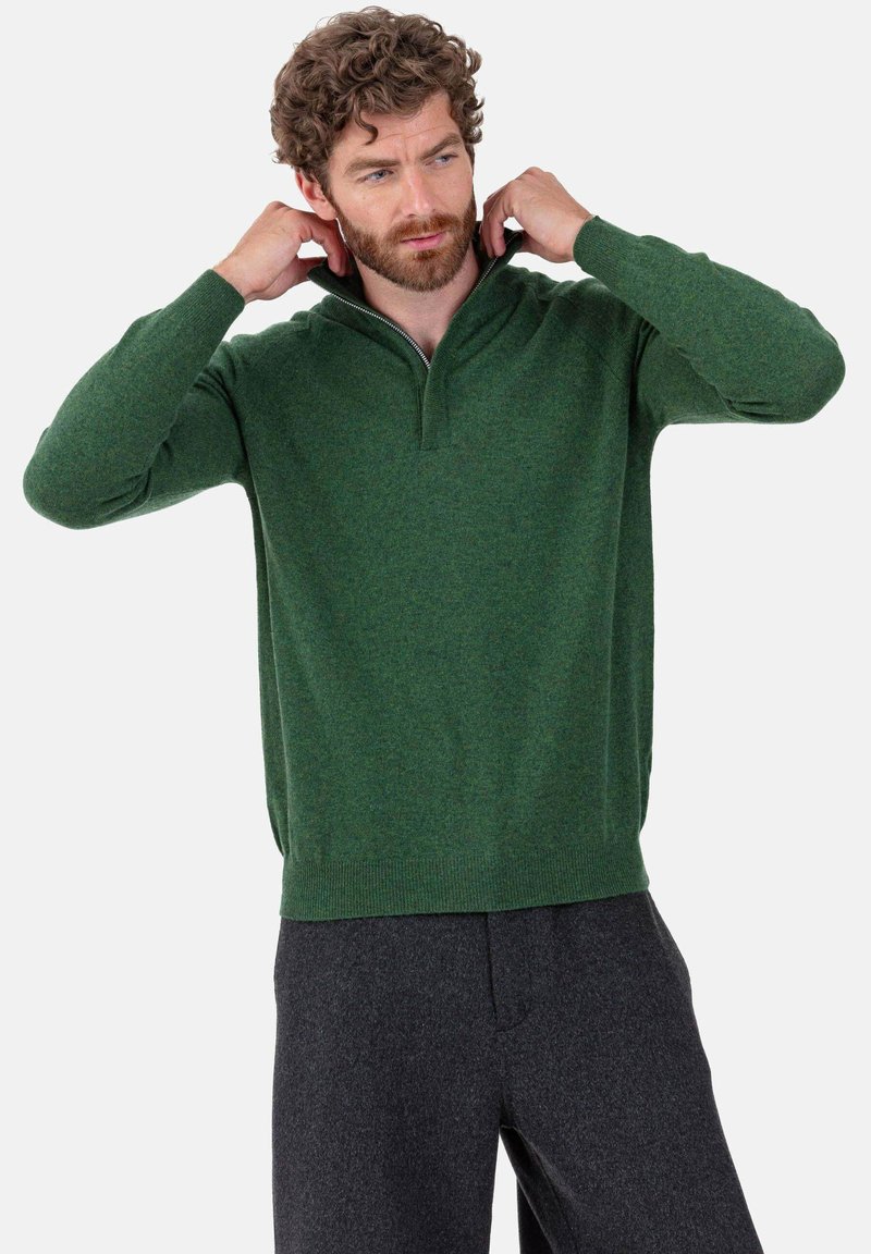 Grüner Strickpullover mit halbem Reißverschluss am Kragen, langen Ärmeln und geripptem Saum. Weiche Textur, lässige Passform, kombiniert mit dunkelgrauen Hosen.