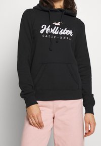 Svart hoodie med snörning, med en vit och rosa logotyp. Materialet verkar mjukt och texturerat. Inkluderar en framficka i känguru-stil.