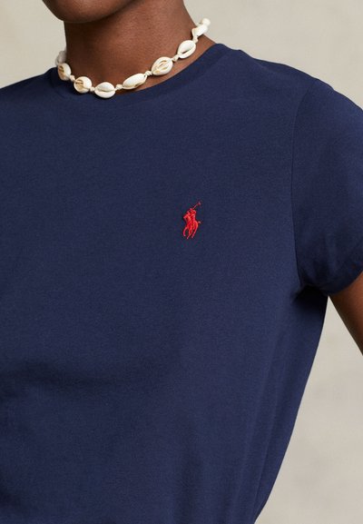 Άτομο που φοράει μπλε σκούρο μπλουζάκι με κόκκινο κεντημένο λογότυπο στο στήθος και λευκό κολιέ από κοχύλια γύρω από τον λαιμό.