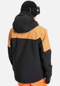 Black and brown waterproof jacket with a hood, featuring a color-block design and textured fabric. Gloves and orange pants visible.