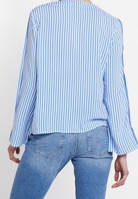 Kobieta ubrana w niebieską i białą pionowo paskowaną koszulę z długim rękawem oraz jasnoniebieskie dżinsy, pokazana z tyłu.