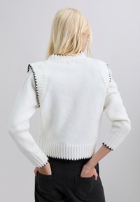 Suéter blanco de punto con dobladillo acanalado y un borde en picos negro en los hombros y las mangas; presenta un cuello alto y un diseño ajustado.