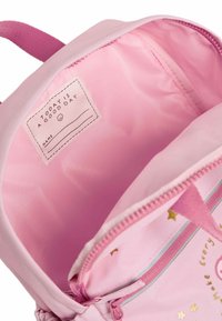 Interiören av en rosa ryggsäck med en slät foder, en etikett med text och små gyllene stjärndetaljer. Dragkedja med en matchande rosa dragplatta.
