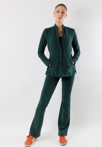 Giacca verde scuro a maniche lunghe con colletto alto e due bottoni, abbinata a pantaloni fluido coordinati e scarpe da ginnastica arancioni.
