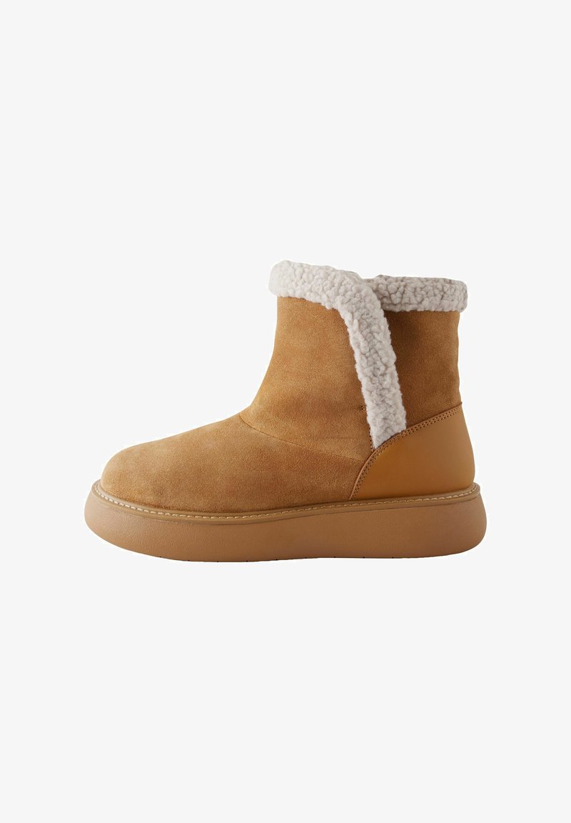Botte à cheville en suède beige avec un col en shearling, bout arrondi et semelle en caoutchouc épaisse. Présente un panneau arrière lisse et une couture contrastante.