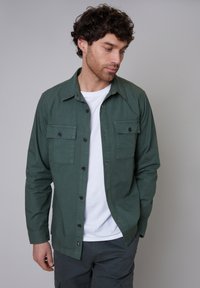 Grüne Hemdbluse aus einem glatten Stoff, mit zwei Brusttaschen, schwarzen Knöpfen und einem klassischen Kragen. Getragen über einem weißen T-Shirt.