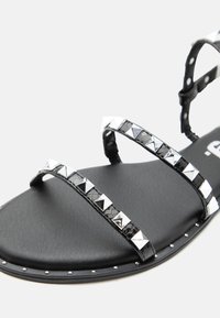 Μαύρο επίπεδο σανδάλι με δύο λουριά διακοσμημένα με ασημένιες πυραμιδοειδείς τρούκ και μικρές ασημένιες στρογγυλές τρούκ κατά μήκος της άκρης της σόλας.