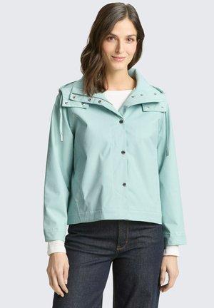 Frau trägt eine hellblaue Kapuzenjacke über einem weißen Hemd und dunklen Jeans, steht vor einem schlichten hellgrauen Hintergrund.