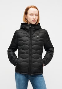 Γυναίκα με κόκκινα μαλλιά που φοράει μαύρο μπουφάν με επένδυση, κουκούλα και φερμουάρ, όρθια με τα χέρια στις τσέπες μπροστά σε απλό φόντο.