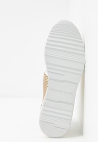 Zapatilla blanca con una suela de goma texturizada, que presenta un panel lateral perforado en beige y un logotipo embossado. Diseño suave y minimalista.