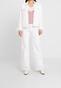 Veste en jean blanche avec des boutons argentés, associée à un haut en tricot rose avec un motif chevron, et un pantalon blanc à jambes larges.