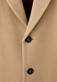 Abrigo beige con tela texturizada, que presenta una solapa con pico y dos botones negros. El forro interior es de color burdeos oscuro.