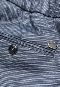 Tissu bleu texturé avec une poche boutonnée et une petite étiquette métallique ovale portant la mention « bugatti » sur le côté droit.