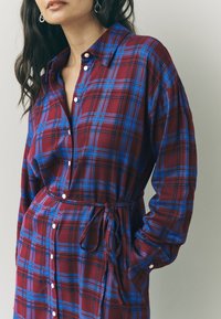 Rød og blå ternet skjortekjole lavet af blødt stof med knaplukning foran, krave, lange ærmer, taljebånd og sidelommer.