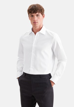 Junger Mann, der ein eng anliegendes weißes Hemd und schwarze Hose trägt, steht mit den Händen in den Taschen vor einem schlichten Hintergrund.