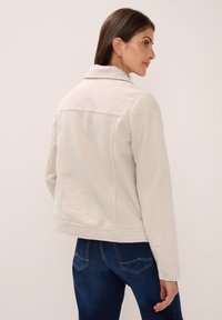Kvinna med fräknig ansikte iklädd en krämfärgad fleecejacka och mörkblå jeans, sedd bakifrån mot en enfärgad bakgrund.