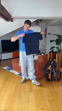 Νέος άνδρας με casual ρούχα κρατάει μια διπλωμένη σκούρα μπλε μπλούζα σε ένα σαλόνι με ένα μηχάνημα κωπηλασίας και καφέ καναπέ.