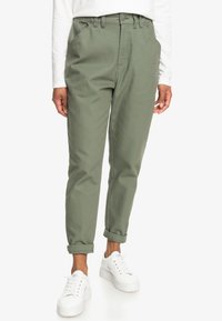Pantalon vert ample à taille haute, jambes fuselées, revers roulés et poches avant. Porté avec des baskets blanches et une chemise à manches longues.