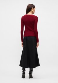 Bordo, raukšlėtas, prigludęs megztinis su ilgomis rankovėmis, derinamas su laisvu juodu midi sijonu. Juodi kelių aukščio aukštakulniai batai pabrėžia ansamblį.